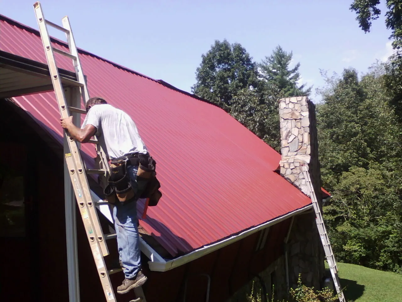 Roofer on a ladder beside a red metal roof and stone chimney — Soffit & Fascia Repair by Roofing Brothers in Rosalie
